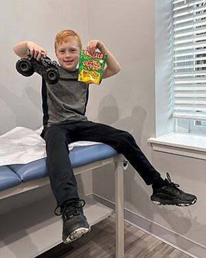 Caleb, an oral immunotherapy patient for peanut allergy, smiles while sitting on a clinic bed with a bag of peanuts and a toy car